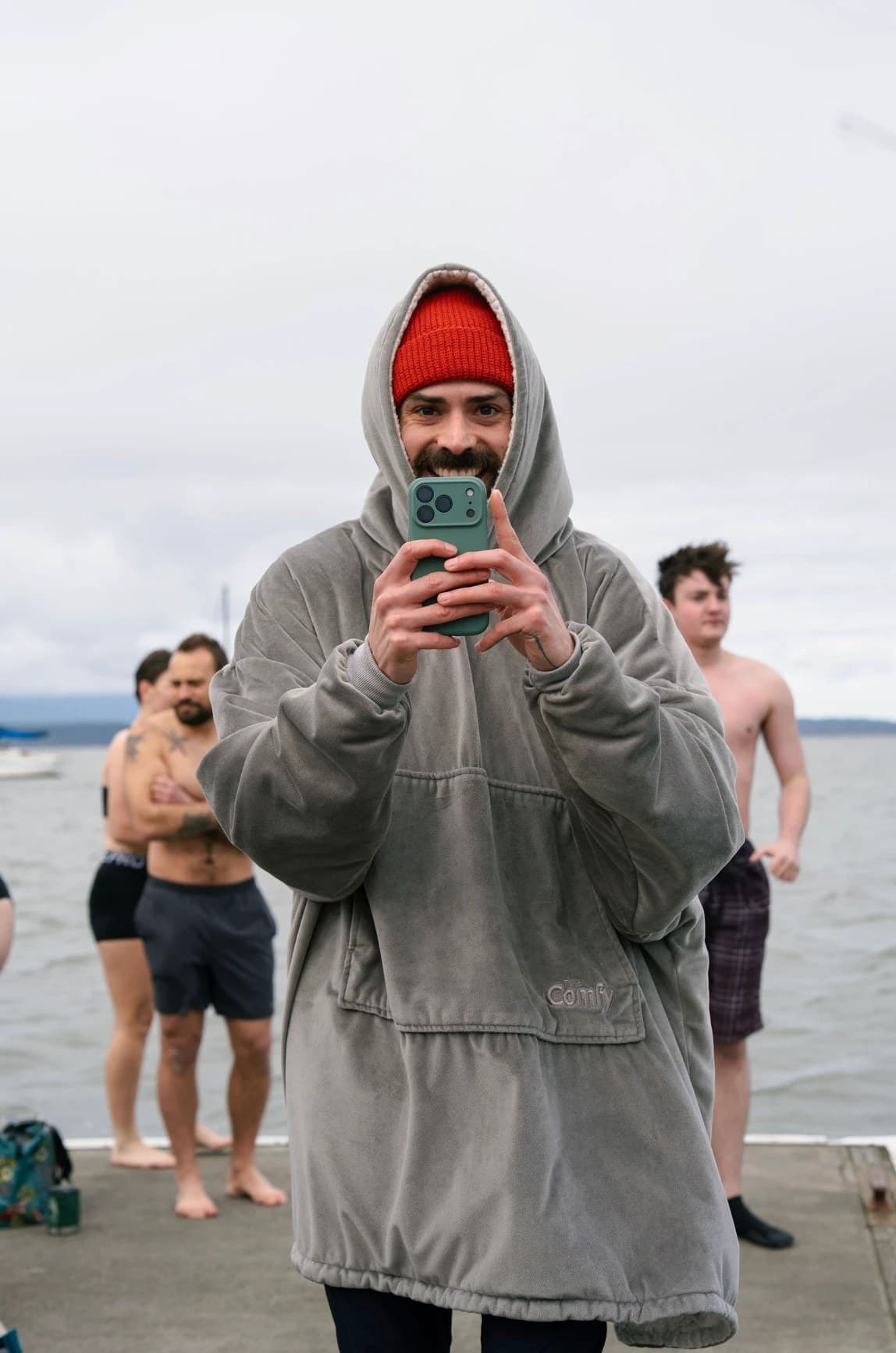 Matt grinning behind his phone on the dock, wrapped in an oversized grey hoodie and a red beanie, with plungers behind him.